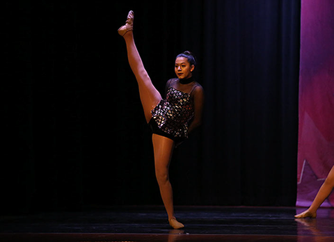 A girl onstage at a dance competition doing a high kick - a grand battement in proprer dance terminology. This is from a jazz dance.