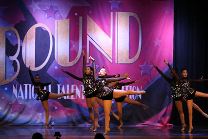A group of girls onstage at Starbound doing arabesques. They are wearing dark glittery costumes. This picture is from a jazz dance.