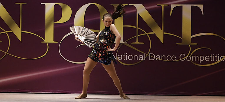 A girl on stage at Starbound National Dance Competition with a dark glittery costume and a fan. This picture is from a jazz dance.