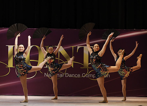 Four girls on stage at OnPoint Dance Competition in "oilslick" costumes and holding fans, doing an attitude. This picture is from a jazz dance.