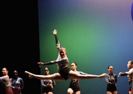 Various girls on stage at Kean University in purple and black costume. One girl is midair doing a perfect leap.